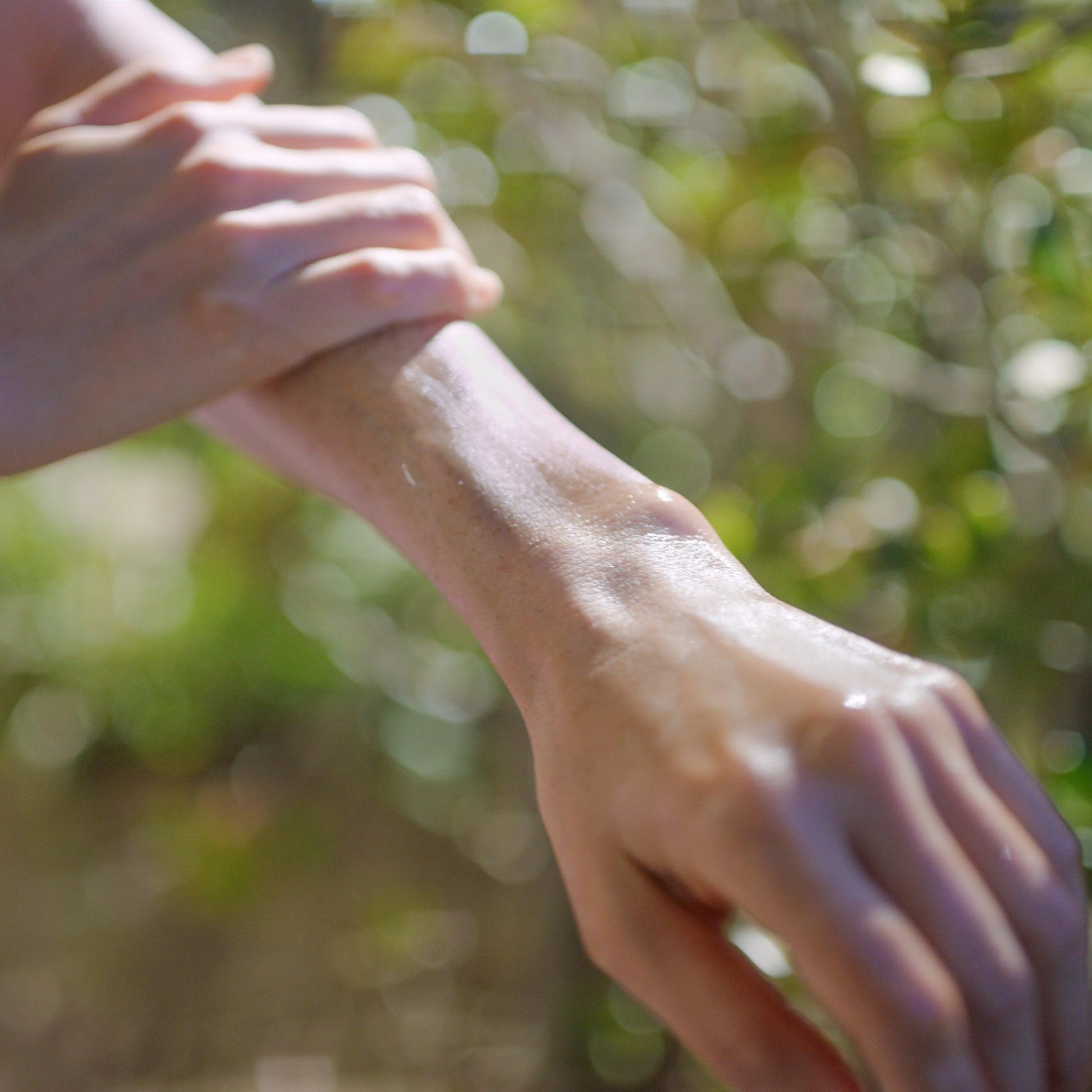 Close-up of a person applying C & The Moon's Malibu Made Glow Oil to their forearm. The visible hand is gently massaging the organic oils into the skin. The background features blurred greenery, evoking an outdoor setting and a clean skincare routine.