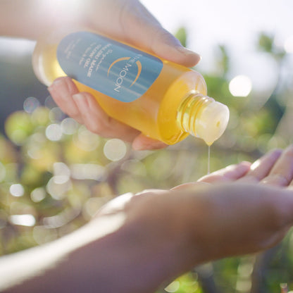 Close-up of hands holding a clear bottle filled with yellow liquid, pouring it onto another open hand below. The label on the bottle reads "C & The Moon Malibu Made Glow Oil". The background is blurred with leafy greenery, suggesting an outdoor setting and a clean skincare routine. The image has a soft, sunlit glow.