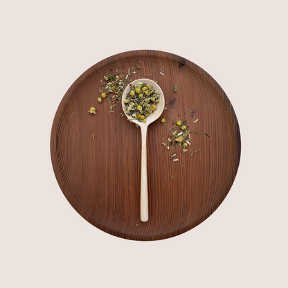 A wooden plate showcases a spoon filled with Moon Canyon Healing's Gut Knowing Tea Blend, an herbal concoction designed for mood support that includes a mix of dried herbs and flowers. Loose herbs are artistically scattered around the spoon on the plate, against a soft, off-white backdrop that enhances the serene ambiance.