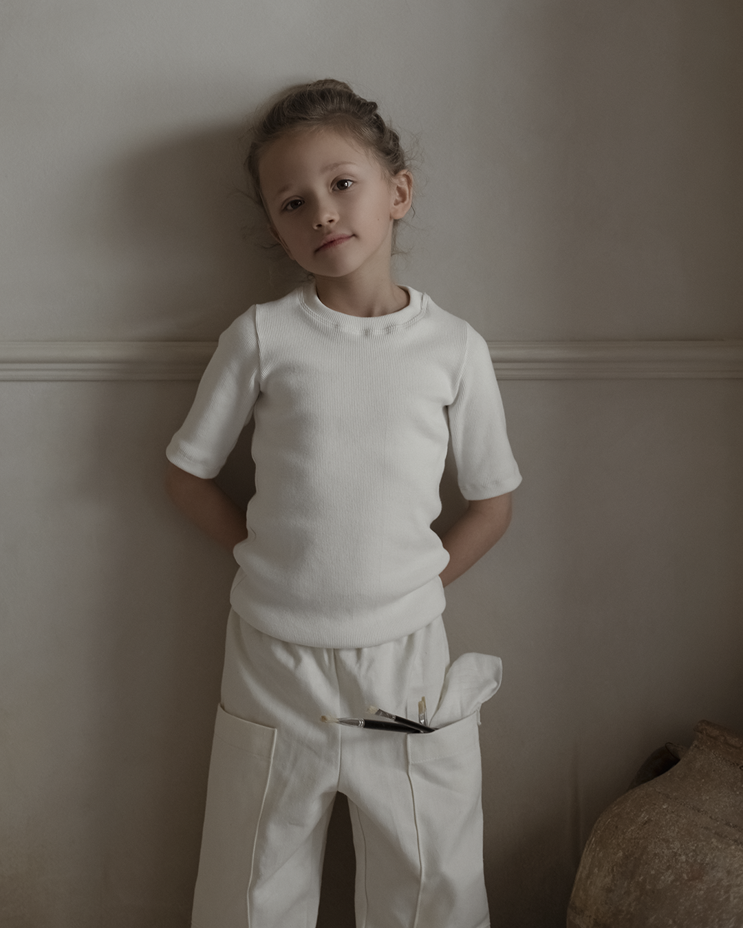 A young child dressed in a cabane childrenswear Classic Top for Baby + Kid and white pants stands against a cream-colored wall, hands resting behind their back. A small paintbrush peeks out from a pocket. The tranquil setting beautifully enhances the clean lines and ethically handmade aesthetic of the ensemble.