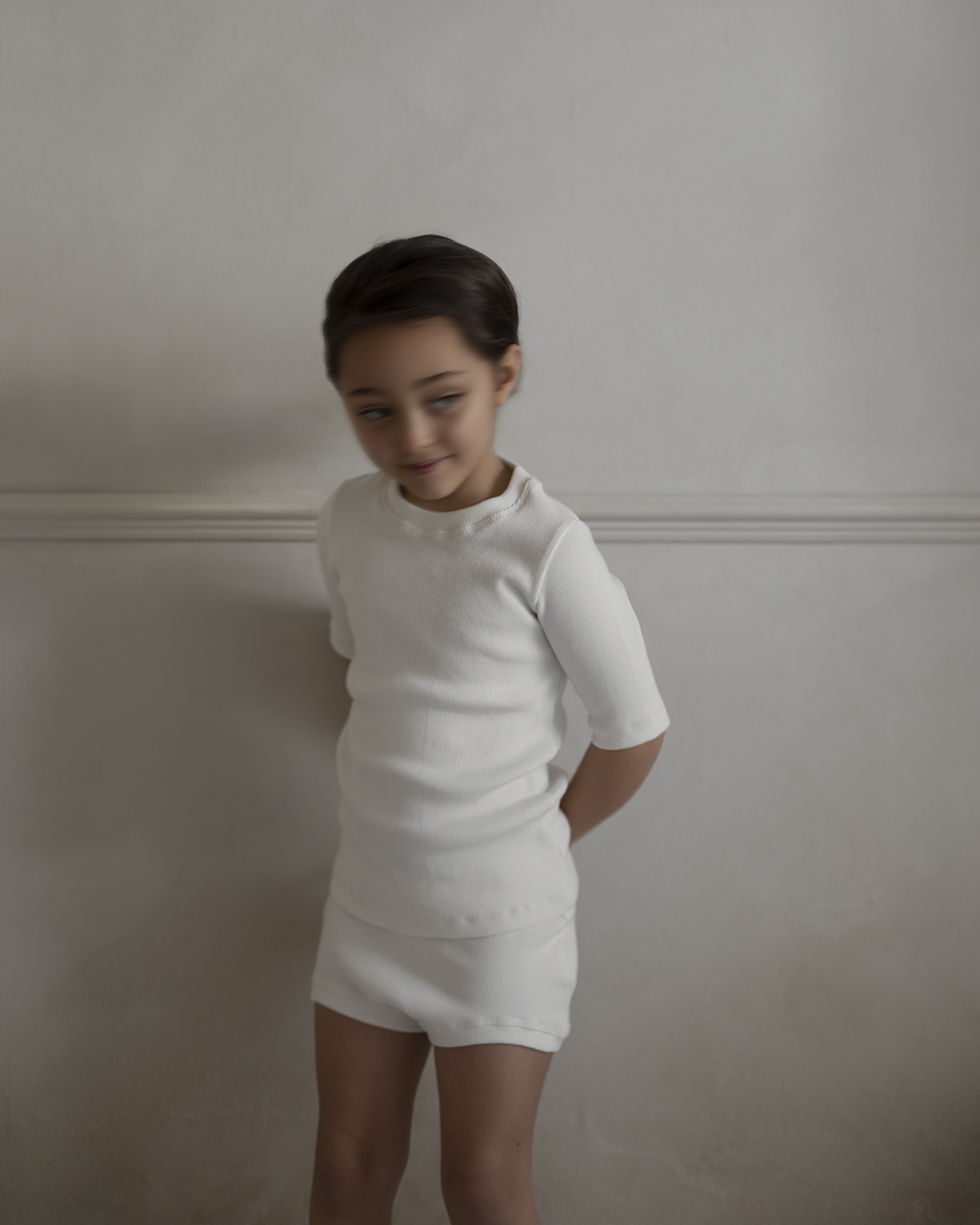 A young child stands against a light beige wall, dressed in the Classic Top for Baby + Kid from cabane childrenswear. The child has short, dark hair and a slight smile while looking away from the camera, creating a soft and minimal atmosphere.