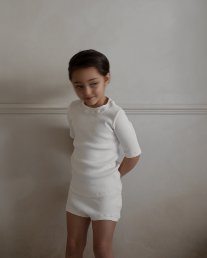 A young child stands against a neutral, slightly textured wall, dressed in a simple white outfit from cabane childrenswear. The child is wearing the CLASSIC TOP baby + kid short-sleeved top paired with matching shorts. They have short dark hair and are looking slightly down and to the side with a subtle smile.