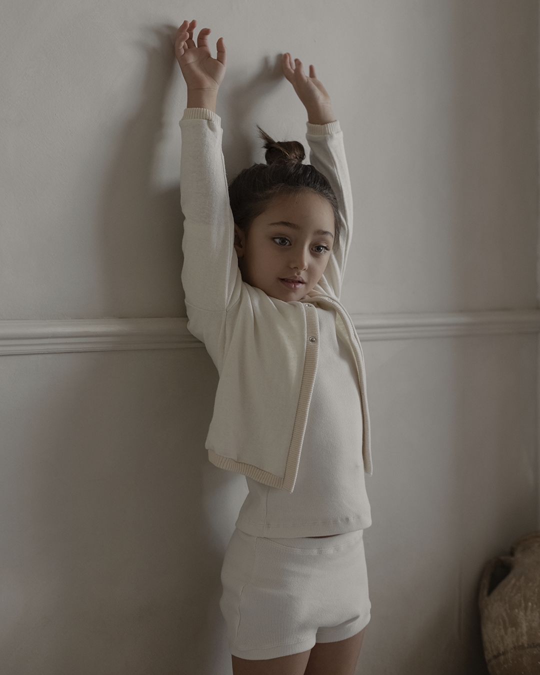 A young child with long dark hair in a bun is stretching with arms up against a gray wall. They are wearing an ethically handmade, light-colored outfit from cabane childrenswear, consisting of the boxy 3-snap Soul Sweater Baby + Kid, top, and shorts. The child stands on a wooden floor next to a beige cushion.