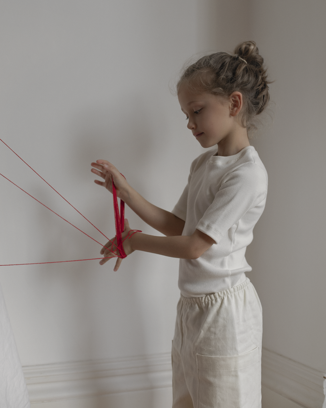 A young girl stands against a white wall, concentrating intensely as she plays with red string wrapped around her fingers like a cat's cradle. She is dressed in an ethically handmade Classic Top for Baby + Kid by cabane childrenswear and light pants, with her hair tied up in a bun.