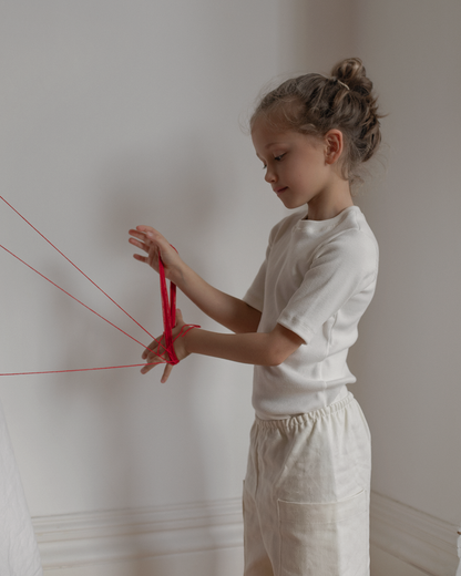 A young girl with her hair in a bun stands against a white wall, intently playing with red string intricately wound around her fingers. She is wearing the CLASSIC TOP baby + kid from cabane childrenswear and light-colored pants, both crafted from ethically handmade materials.