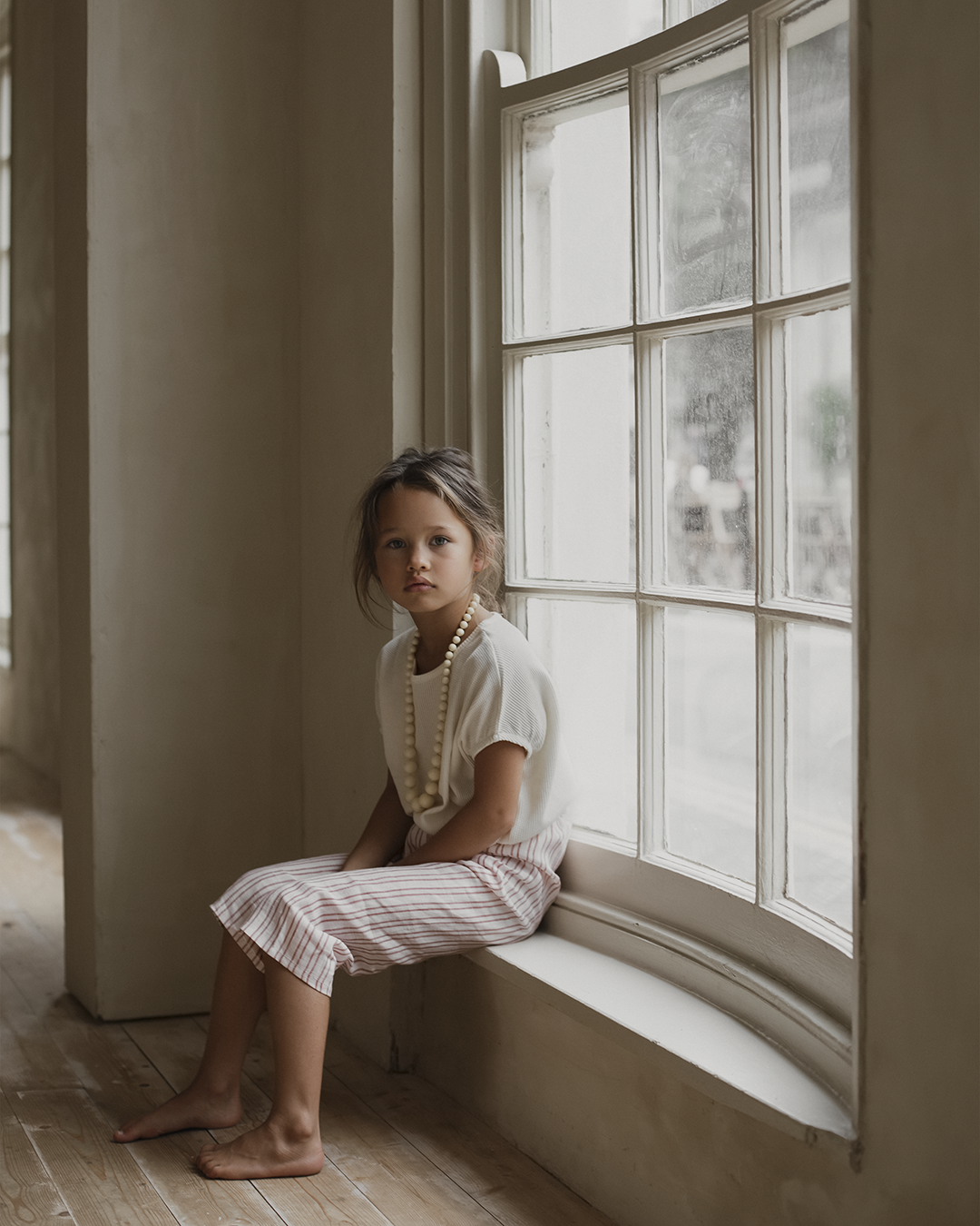 A young girl with tousled hair sits on a wide windowsill, wearing a white top, the PER SE PANT loom linen from cabane childrenswear, and a large beaded necklace. She gazes thoughtfully out the window, with light filtering through and illuminating the room. The decor is minimalistic, with wooden floors and ethically made accents.