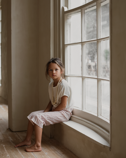 A young girl with tousled hair sits on a wide windowsill, wearing a white top, the PER SE PANT loom linen from cabane childrenswear, and a large beaded necklace. She gazes thoughtfully out the window, with light filtering through and illuminating the room. The decor is minimalistic, with wooden floors and ethically made accents.