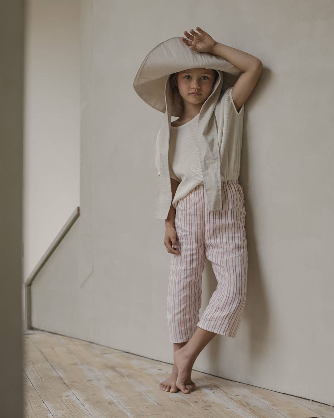 A young child stands barefoot on a wooden floor, leaning against a light-colored wall. Wearing a wide-brimmed hat, an off-white shirt, and handmade white and pink striped PER SE PANT loom linen from cabane childrenswear, the child raises one arm and gazes forward with a serious expression.