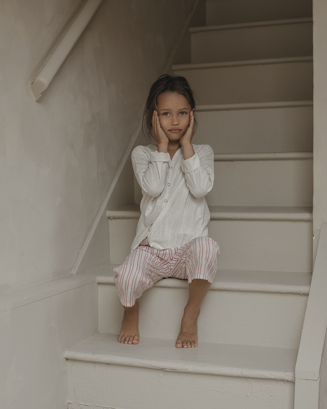 A child with long dark hair sits on white stairs indoors, wearing a light-colored long-sleeve shirt and pink-striped PER SE PANT loom linen by cabane childrenswear. The child holds their face with both hands and looks directly at the camera with a neutral expression.