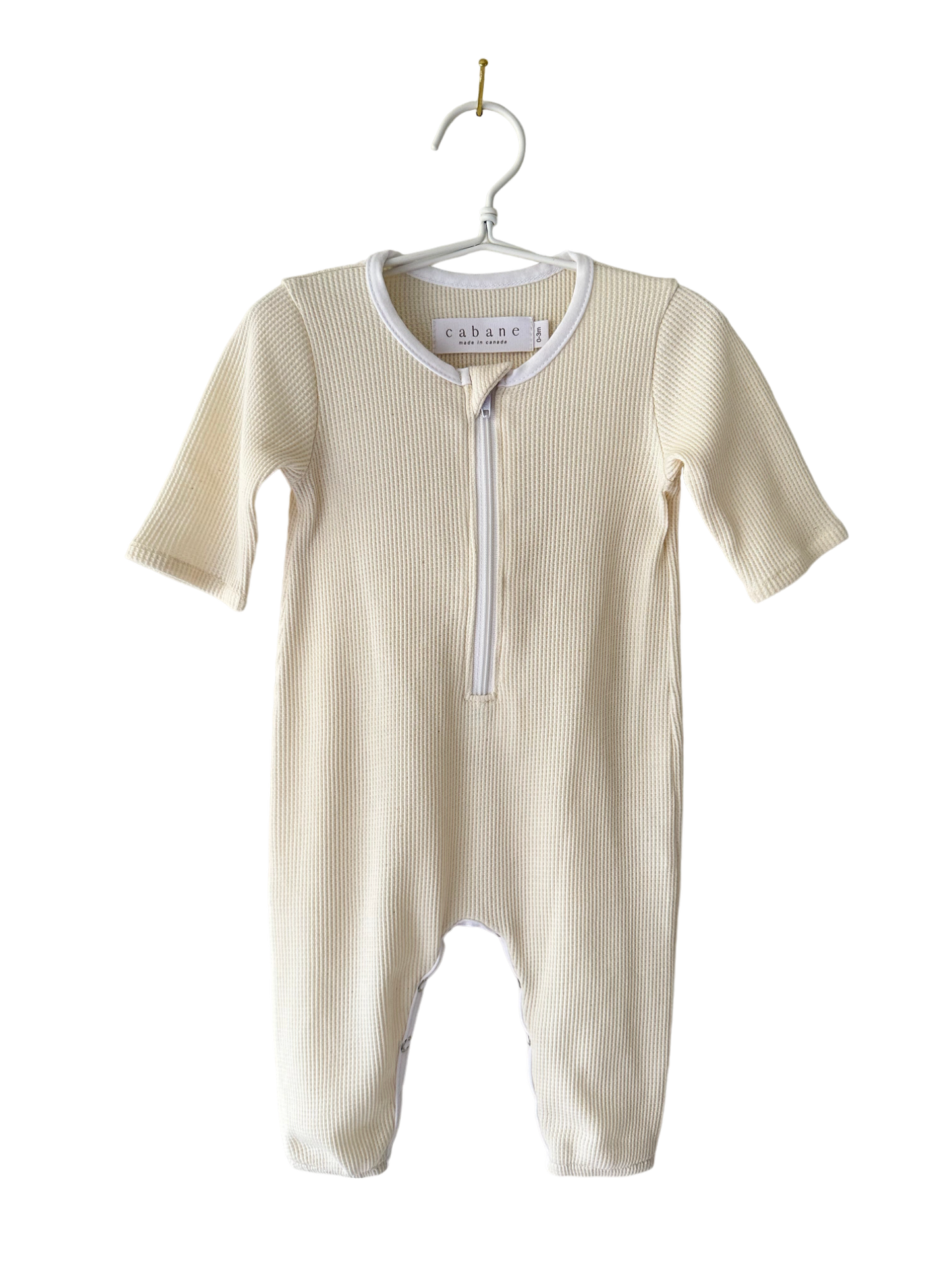 A cream-colored baby onesie from cabane childrenswear, known as THE ORB SUIT WAFFLE baby + kid, with long sleeves and legs, is hanging on a white clothes hanger. The onesie features a convenient zipper running from the neck down to the crotch for easy diaper changes. Ethically handmade, its ribbed texture makes it an ideal seasonless unisex outfit.