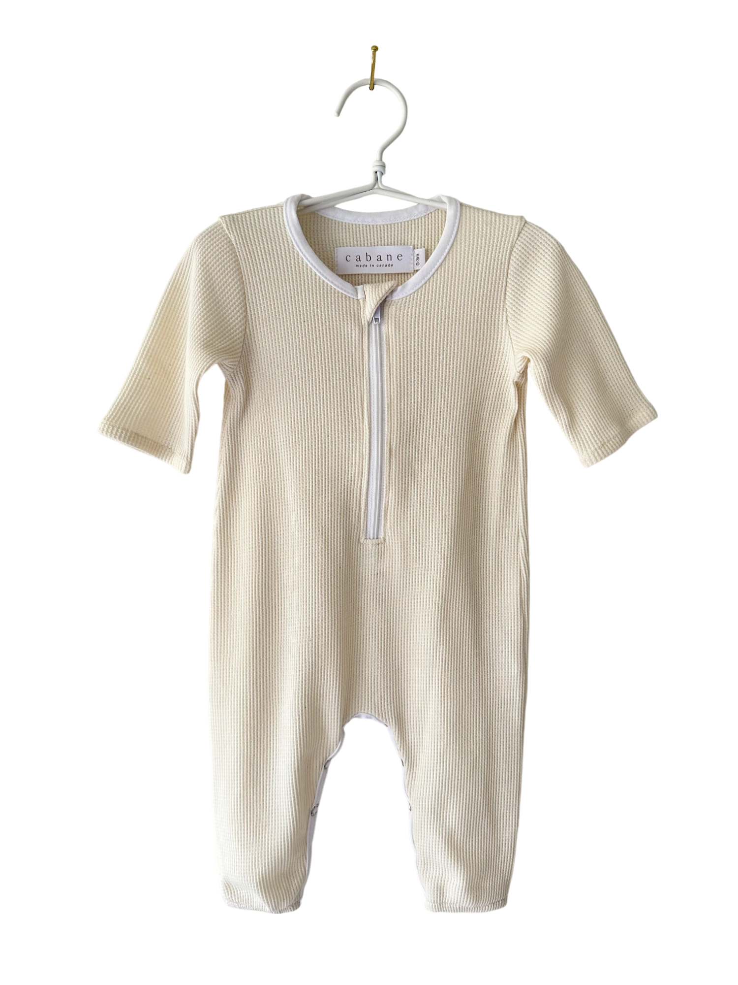 A cream-colored baby onesie from cabane childrenswear, known as THE ORB SUIT WAFFLE baby + kid, with long sleeves and legs, is hanging on a white clothes hanger. The onesie features a convenient zipper running from the neck down to the crotch for easy diaper changes. Ethically handmade, its ribbed texture makes it an ideal seasonless unisex outfit.