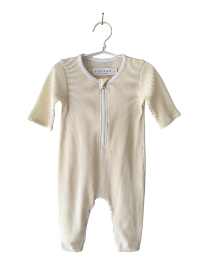 A cream-colored baby onesie from cabane childrenswear, known as THE ORB SUIT WAFFLE baby + kid, with long sleeves and legs, is hanging on a white clothes hanger. The onesie features a convenient zipper running from the neck down to the crotch for easy diaper changes. Ethically handmade, its ribbed texture makes it an ideal seasonless unisex outfit.