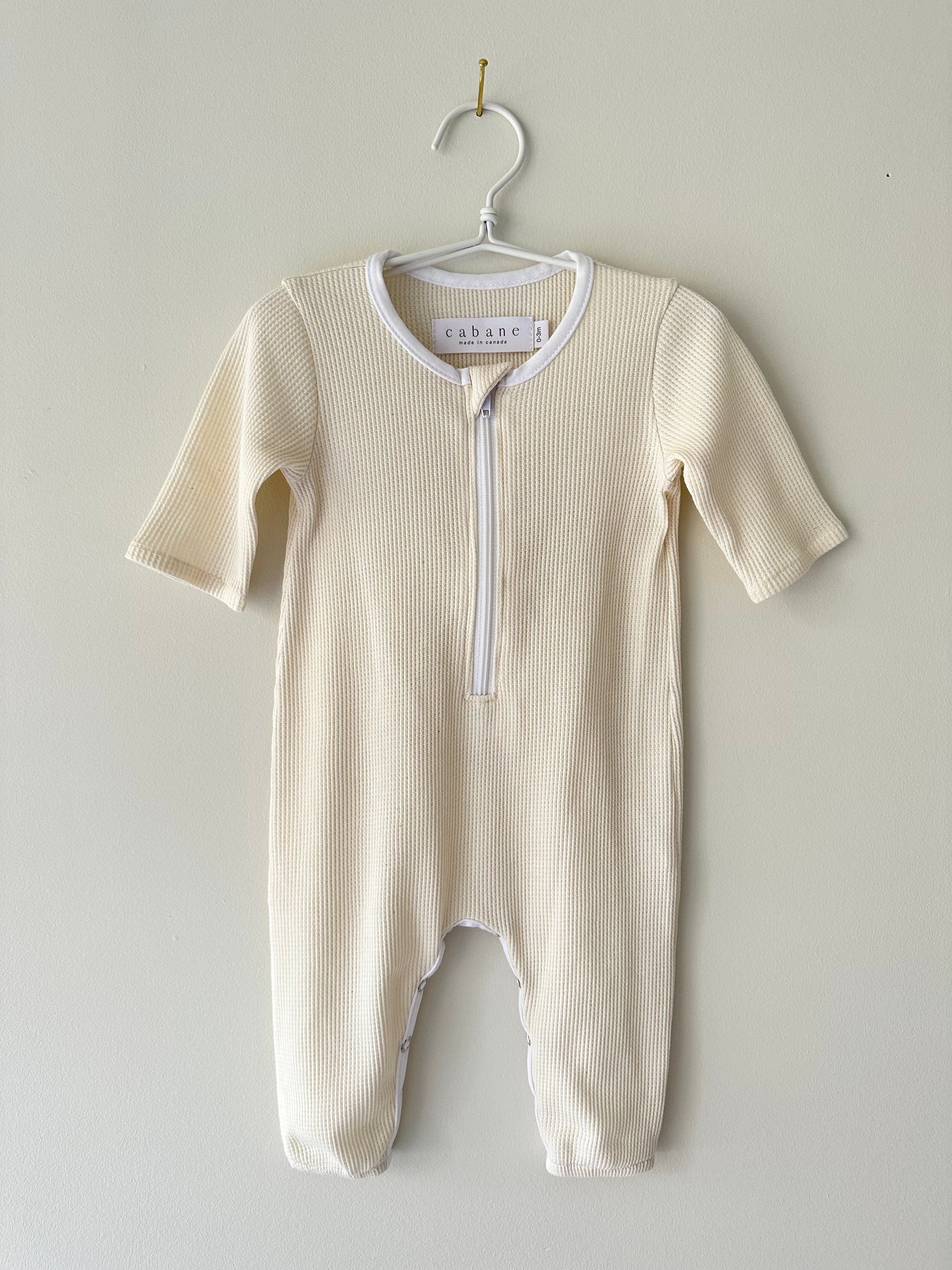 A beige, ribbed baby onesie from cabane childrenswear hangs on a white hanger against a light wall. This seasonless unisex outfit, named THE ORB SUIT WAFFLE baby + kid, features long sleeves, long pants, and a zippered front closure for easy diaper changes. The tag inside reads "cabane," hinting at its ethically handmade origin.