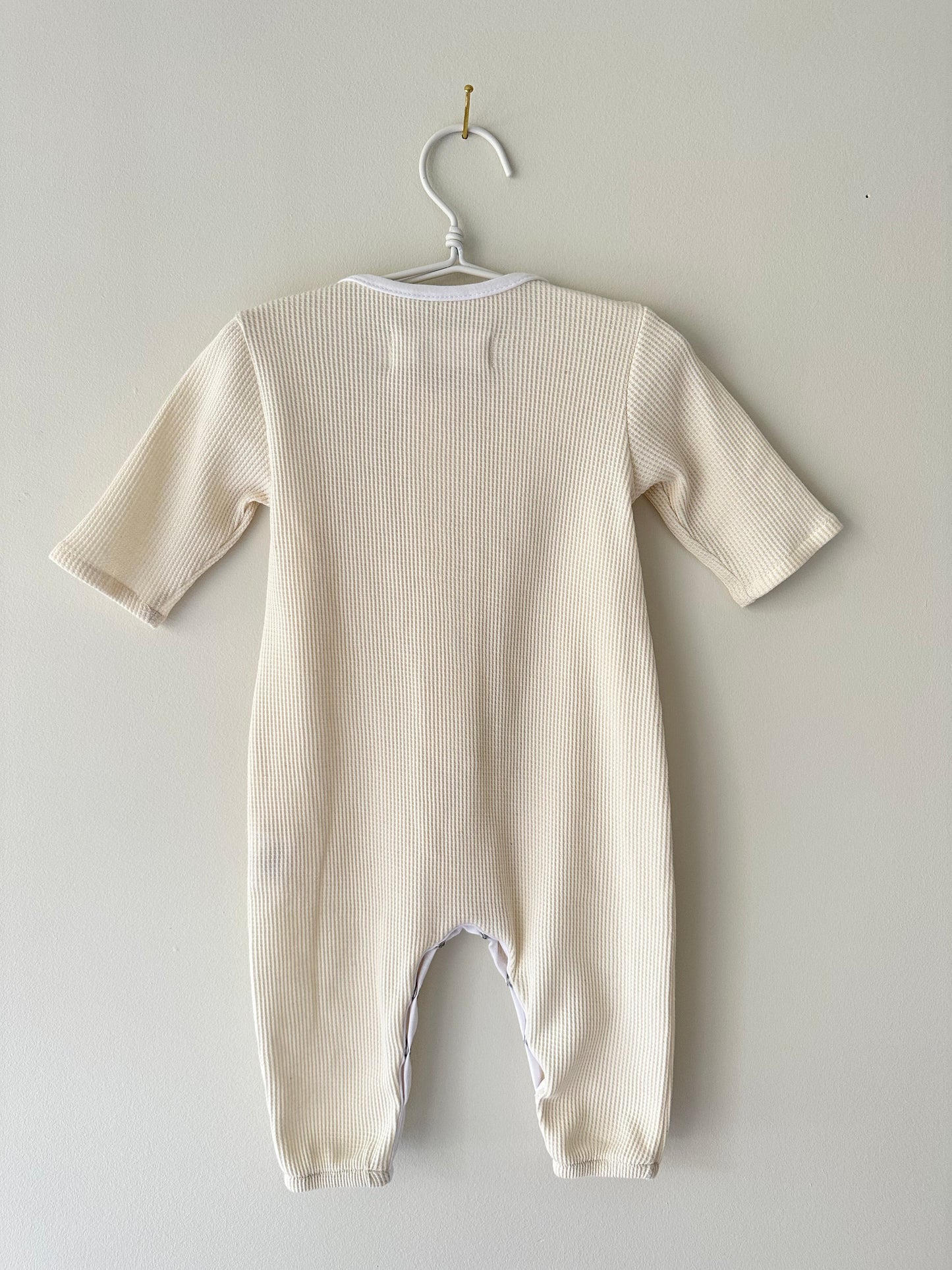 THE ORB SUIT WAFFLE baby + kid from cabane childrenswear is displayed on a white hanger against a light gray wall. The beige, long-sleeve onesie features soft and cozy ribbed fabric, making it an ideal seasonless unisex outfit for your little one. Ethically handmade, this simple design guarantees comfort and ease of wear for both parent and baby.