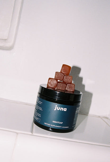 A dark-colored container labeled "Juna" and "Nightcap Sleep Gummies: 50 Gummies" rests on a tiled surface. The open container has a stack of brown gummies piled on top, infused with functional botanicals for restorative sleep. The background is a plain, off-white wall.