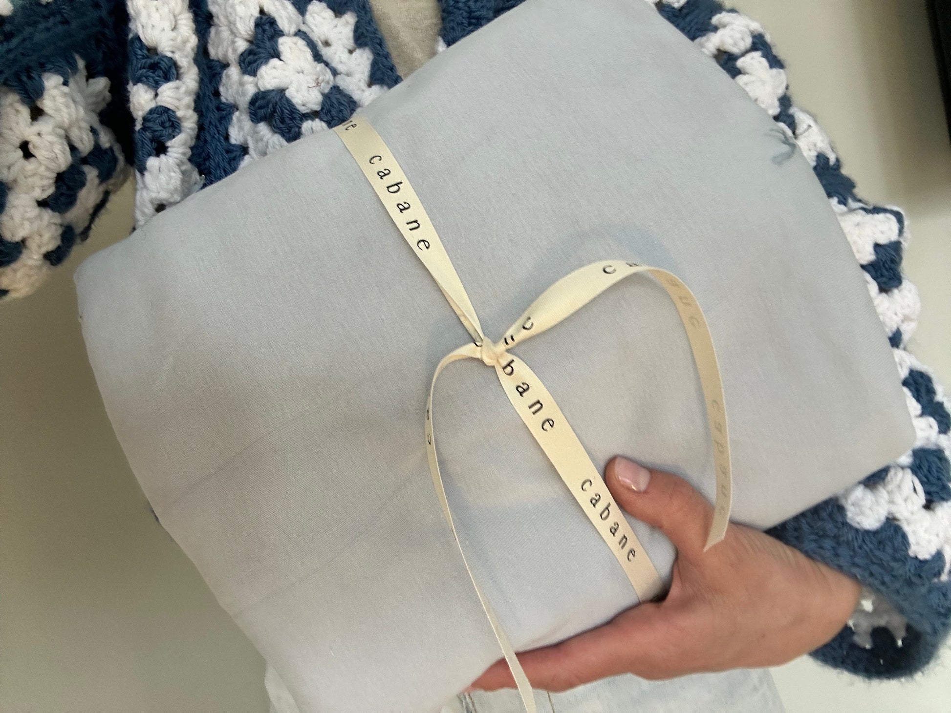 A hand holds a neatly folded light gray fabric wrapped with a cream "cabane" ribbon. In the background, a blue and white crocheted item complements the artisanal touch, evoking the pre-order BABY BLANKET, indigo by cabane childrenswear.