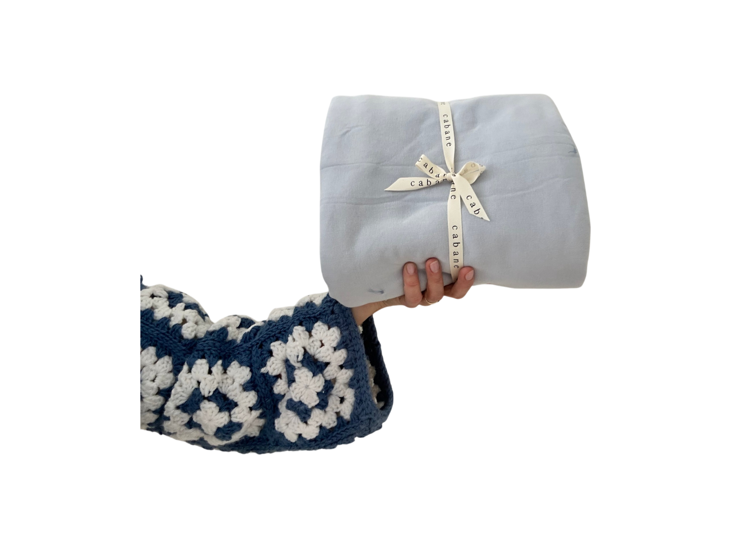 A hand in a blue and white crocheted sleeve holds a neatly folded light gray baby playmat secured with a "cabane childrenswear" ribbon against a black background.