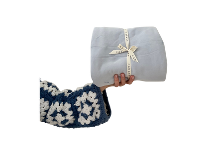 A hand in a blue and white crocheted sleeve holds a neatly folded light gray baby playmat secured with a "cabane childrenswear" ribbon against a black background.