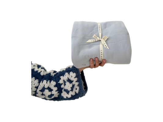 A hand in a blue and white crocheted sleeve holds a neatly folded light gray baby playmat secured with a "cabane childrenswear" ribbon against a black background.