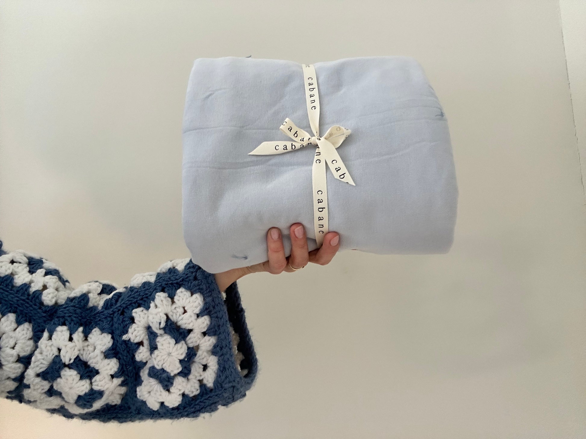 A person holds a neatly folded light blue "pre-order BABY BLANKET, indigo" by cabane childrenswear, resembling a hand-quilted blanket. They're wearing a blue and white geometric crochet sweater against a plain light-colored wall.