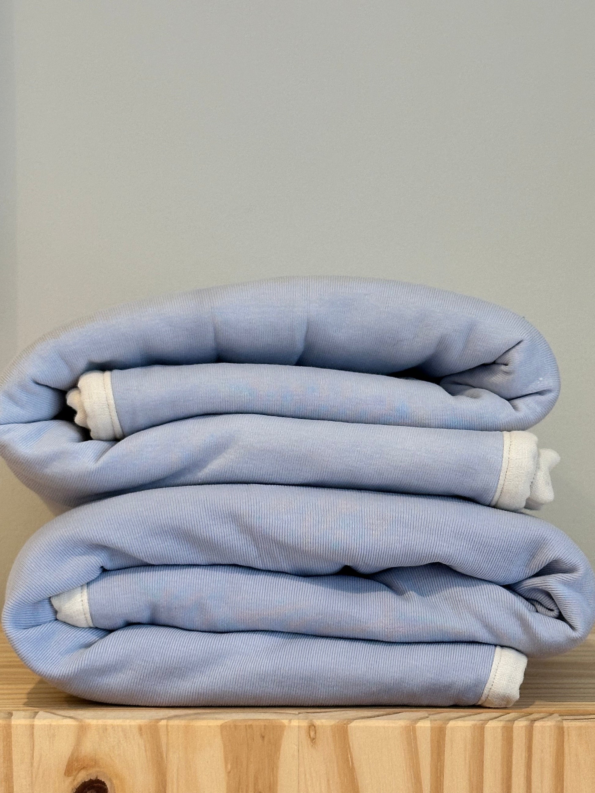 A neatly folded stack of light blue bed linens with white edges, accompanied by a pre-order BABY BLANKET in indigo from cabane childrenswear, sits on a wooden surface against a light gray wall.