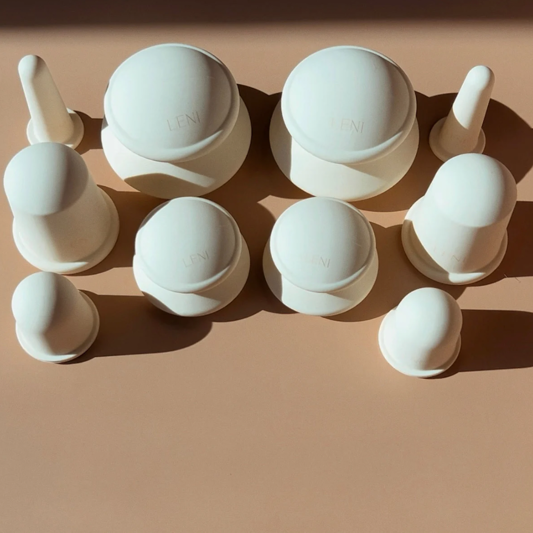 A collection of variously sized and shaped white silicone suction cups from The Face & Body Cupping Set by LENI are evenly spaced on a light brown surface. Arranged in a neat grid pattern, they cast subtle shadows suggesting a light source from the top right.