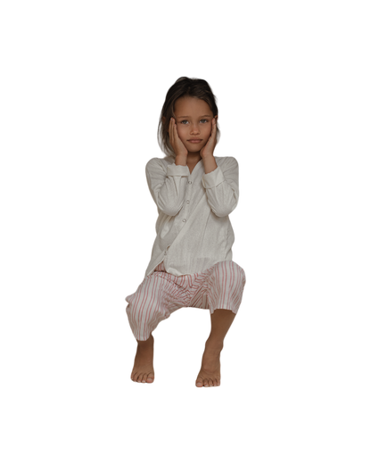 A young child with long hair, wearing the TAKE CARE TOP raw silk long-sleeve shirt by Cabane Childrenswear and white and pink striped pants, is crouching down with their hands on their cheeks against a black background.