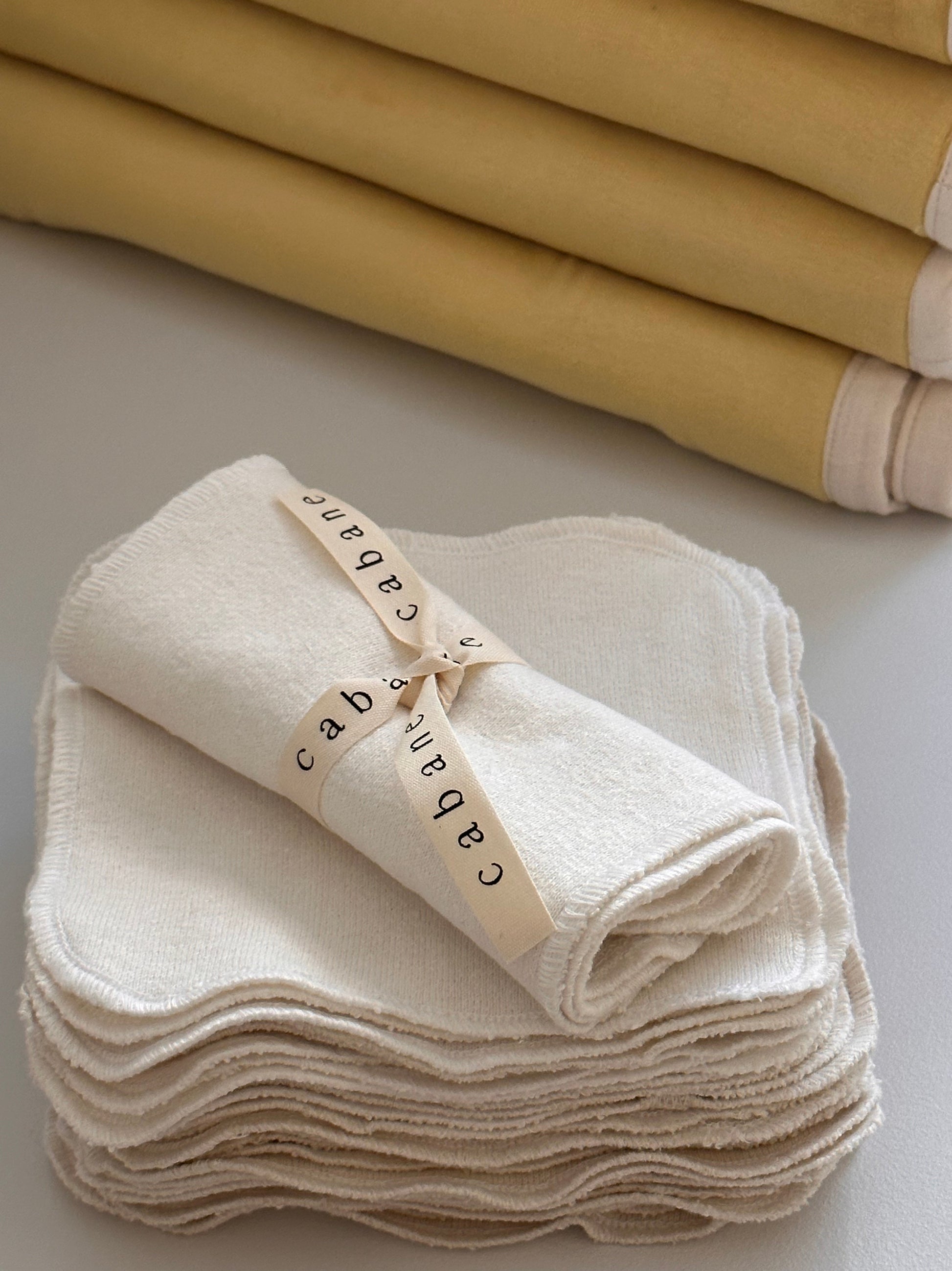 A neatly stacked pile of cream-colored, undyed organic cotton cloths is placed on a table. On top of the pile, a single cloth is rolled up and secured with a ribbon that reads "cabane childrenswear." In the background, there is another stack of folded yellow washcloths from the Organic Washcloth Set of 4.