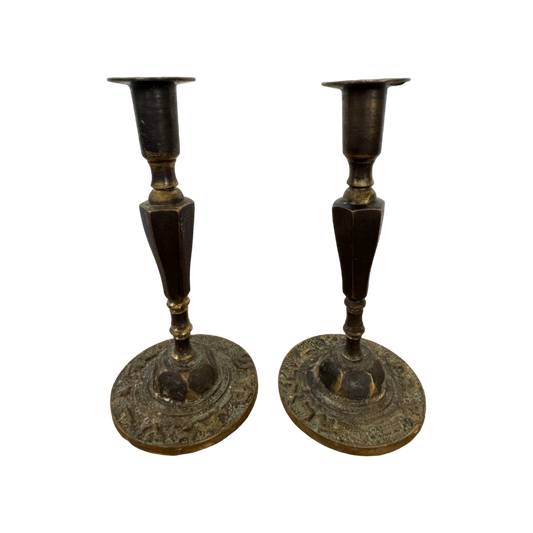 A pair of Shoppe Maison Vintage Shabbat Candle Holders with tall, tapered shafts and wide, circular bases featuring intricate etched designs, set against a black background. These brass candlesticks show signs of tarnish, adding to their antique appearance.