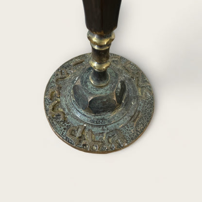 A close-up view of the base of an ornate metal object, which appears to be a Vintage Shabbat Candle Holder from Shoppe Maison. The base is adorned with intricate engravings and has a weathered, slightly tarnished patina, featuring decorative Hebrew lettering around its circular bottom.