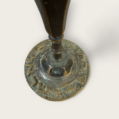 A close-up of the base of an ornate metal object, possibly a candlestick. The base is round with intricate engravings along the edge and a slightly weathered patina. The central stem appears cylindrical and tapers as it extends upward, resembling the Vintage Shabbat Candle Holders by Shoppe Maison.