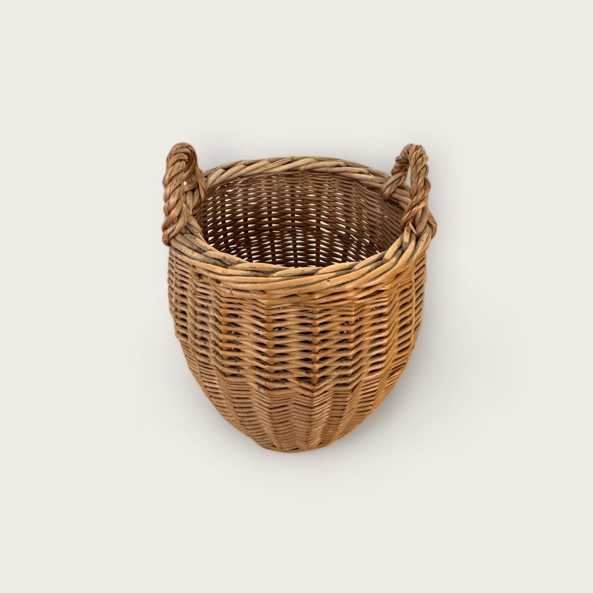 Product Description: The Small Vintage Basket with Handles from Shoppe Maison is a round, woven wicker basket featuring two thick handles on opposite sides. This light brown basket showcases tight and even weaving, exuding a natural rustic charm. It stands on a flat base and is displayed empty against a plain white background. Perfect for homewares enthusiasts.