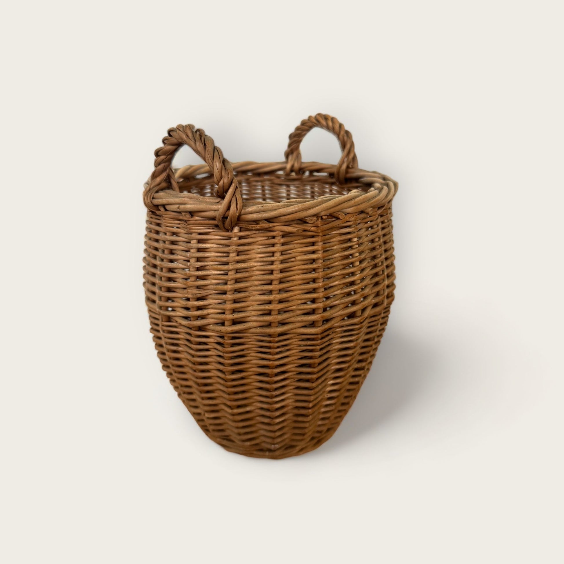 Introducing the Small Vintage Basket with Handles from Shoppe Maison. This handwoven wicker basket features a rounded bottom and two twisted handles on top. With its natural brown color and intricate weaving pattern, it boasts a rustic and artisanal appearance. Displayed against a plain light background, this charming vintage basket adds timeless elegance to any space.