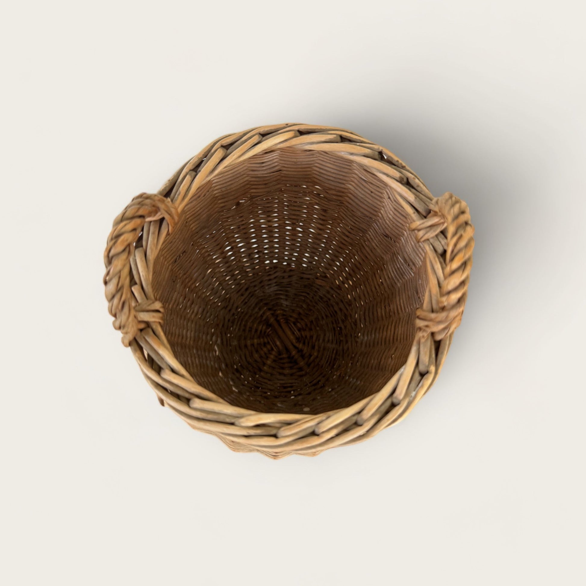 A top-down view of the Small Vintage Basket with Handles from Shoppe Maison reveals its empty, round wicker form with two loop handles on the sides. The light brown woven material exudes charm, making it an ideal vintage basket for any homewares collection. This elegant piece is a perfect addition to Shoppe Maison's curated selection, set against a plain white background.