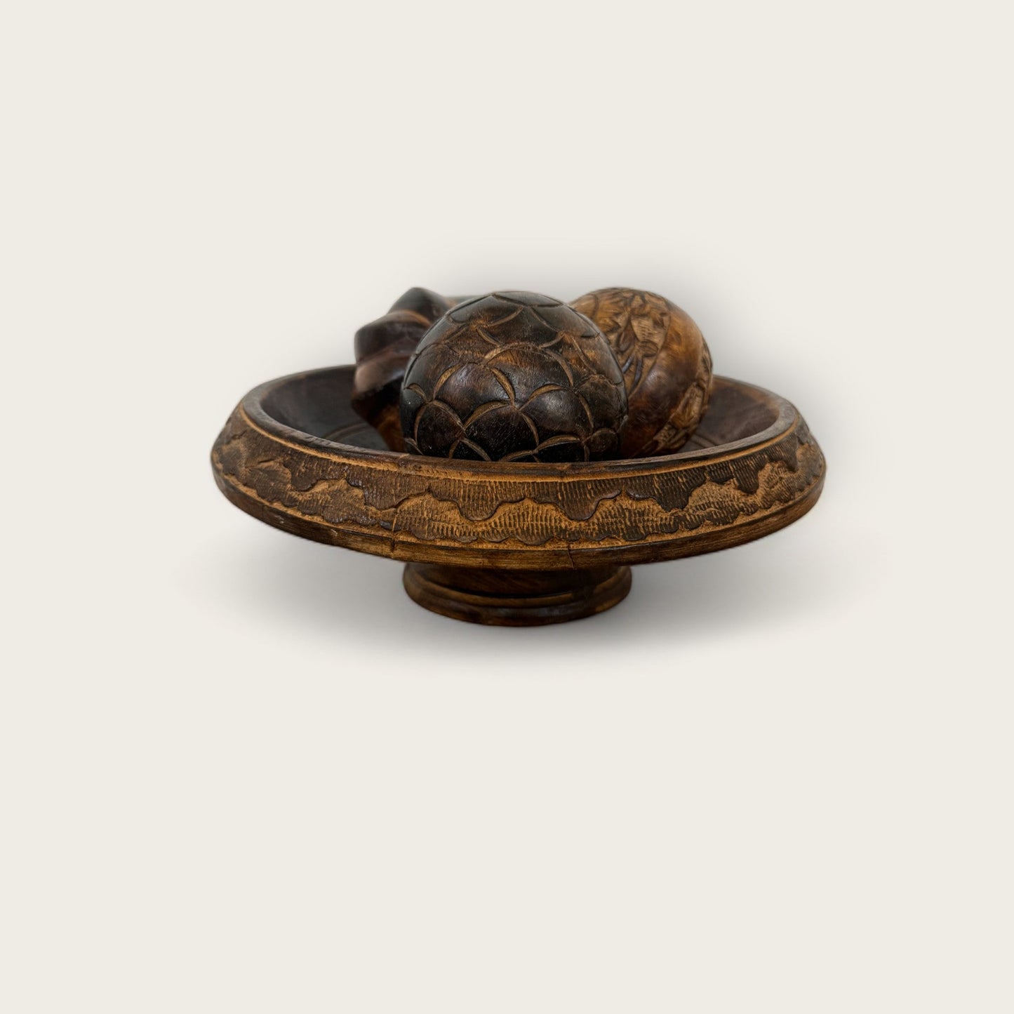 An ornately engraved wooden bowl featuring floral patterns gently holds two intricately carved balls with geometric designs. This elegant ensemble is perched on a Carved Wood Pedestal from Shoppe Maison, set against a simple white backdrop.