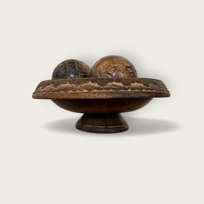 A Shoppe Maison Carved Wood Pedestal, featuring intricate carvings around its rim, holds two hand-carved wooden spheres. The pedestal and spheres are displayed against a plain, light-colored background.