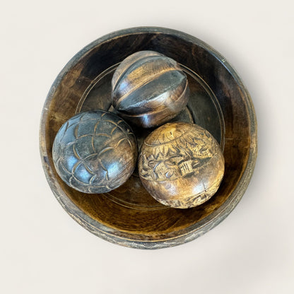 A Carved Wood Pedestal by Shoppe Maison holds a wooden bowl with three intricately hand-carved wooden balls. The dark wood, adorned with natural grain patterns and detailed carvings, is beautifully contrasted against a plain, light-colored background, highlighting the exquisite craftsmanship.