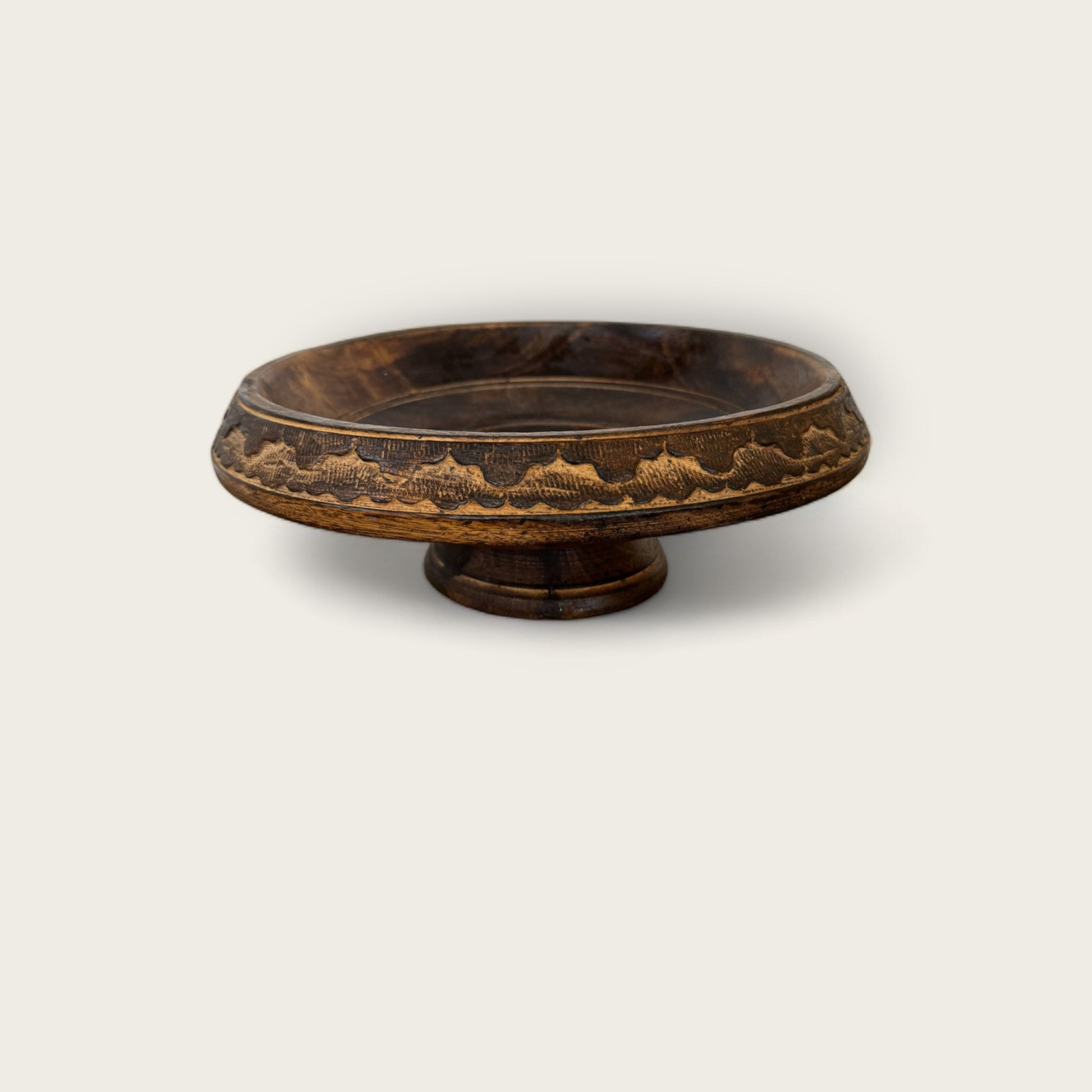 A photograph of the Shoppe Maison Carved Wood Pedestal, a vintage hand-carved bowl featuring intricate patterns around its rim. The pedestal has a round base that raises it off the ground and expands into a wider, shallow bowl section. The rich, dark finish accentuates the detailed craftsmanship typical of fine vintage goods.