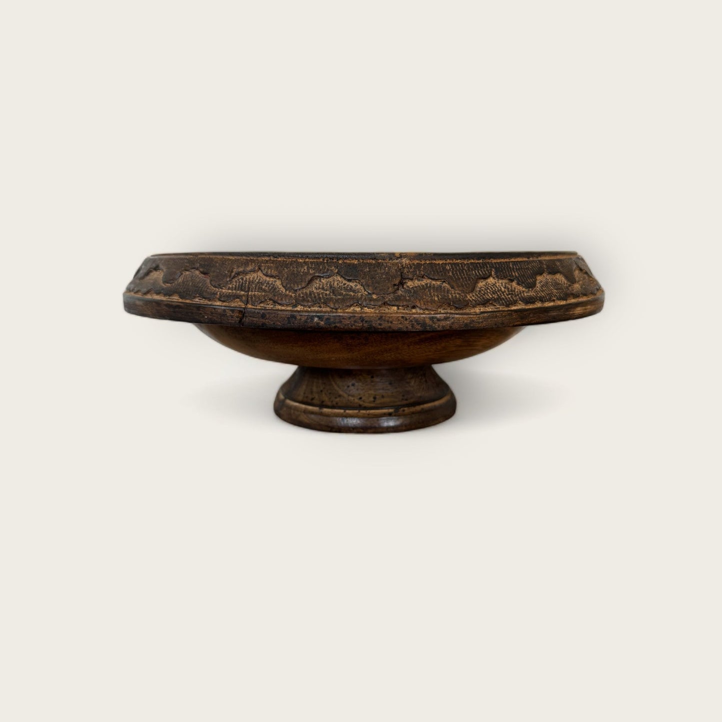 The Shoppe Maison Carved Wood Pedestal is a vintage dark wooden bowl with a flat, circular base and a wide, shallow basin. Its rim features an intricate hand-carved pattern, giving it a rustic and ornate appearance. The plain, light background highlights the pedestal's exquisite details.