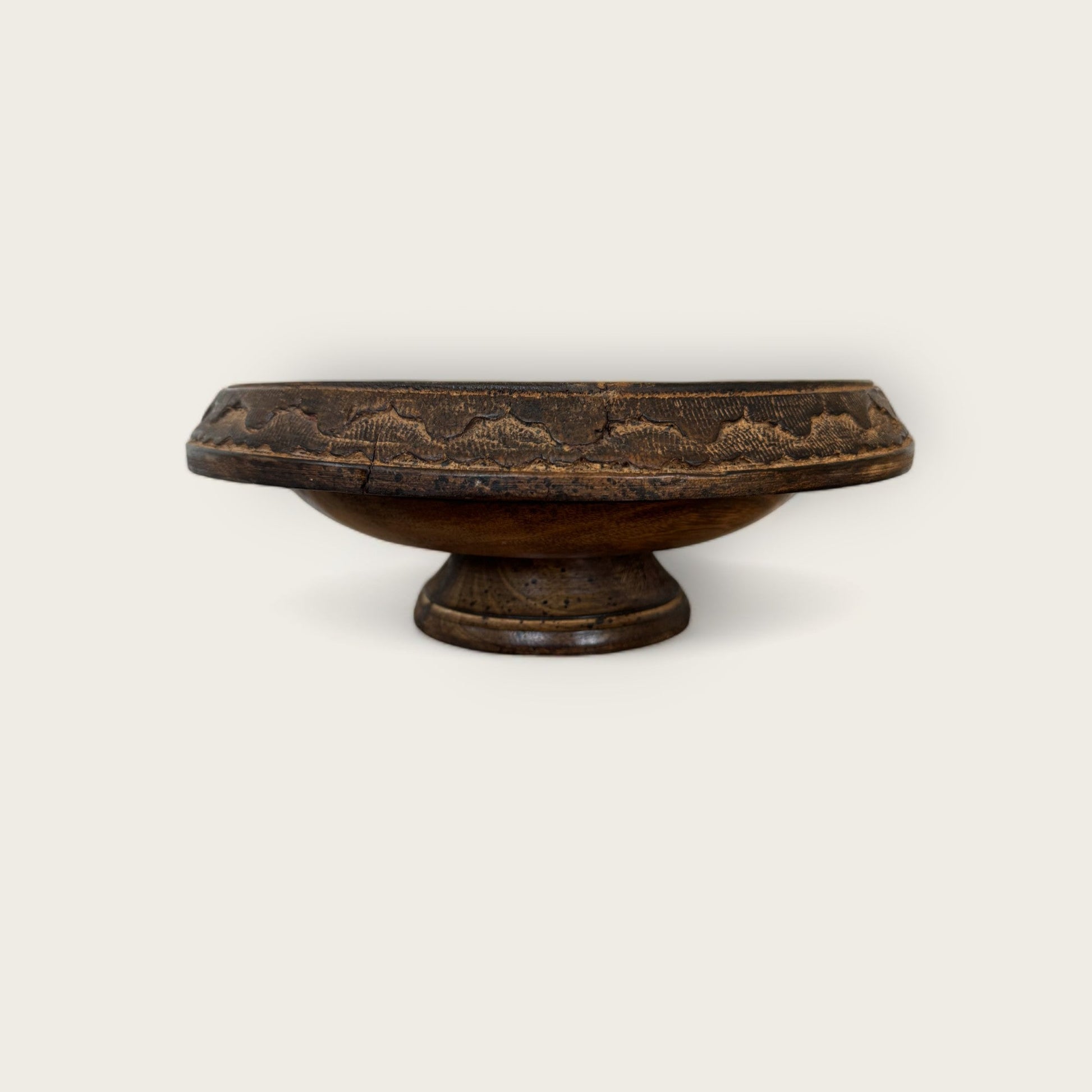 The Shoppe Maison Carved Wood Pedestal is a vintage dark wooden bowl with a flat, circular base and a wide, shallow basin. Its rim features an intricate hand-carved pattern, giving it a rustic and ornate appearance. The plain, light background highlights the pedestal's exquisite details.