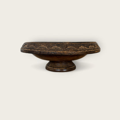 The Shoppe Maison Carved Wood Pedestal is a vintage dark wooden bowl with a flat, circular base and a wide, shallow basin. Its rim features an intricate hand-carved pattern, giving it a rustic and ornate appearance. The plain, light background highlights the pedestal's exquisite details.