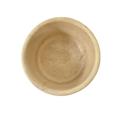 An empty round beige ceramic bowl with a slightly rough texture and visible cracks inside, viewed from above against a solid black background. Resembling the Vintage Small Stoneware Crock from Shoppe Maison, the bowl appears aged and weathered.