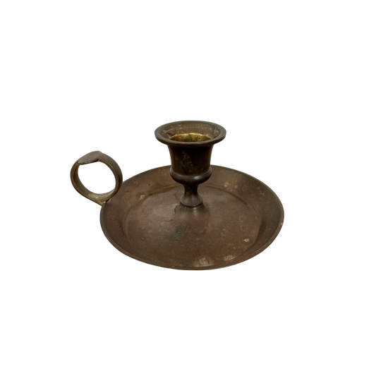 The Shoppe Maison Brass Candle Holder II, featuring a round base, drip tray, and finger ring handle, is set against a black background. Its signs of aging and tarnish enhance its antique charm.