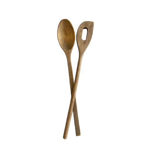 The "Pair of Vintage Wood Spoons" from Shoppe Maison are displayed on a black background with their handles crisscrossed at the bottom; one is a solid spoon while the other is a spatula featuring an elongated oval-shaped hole in the center.