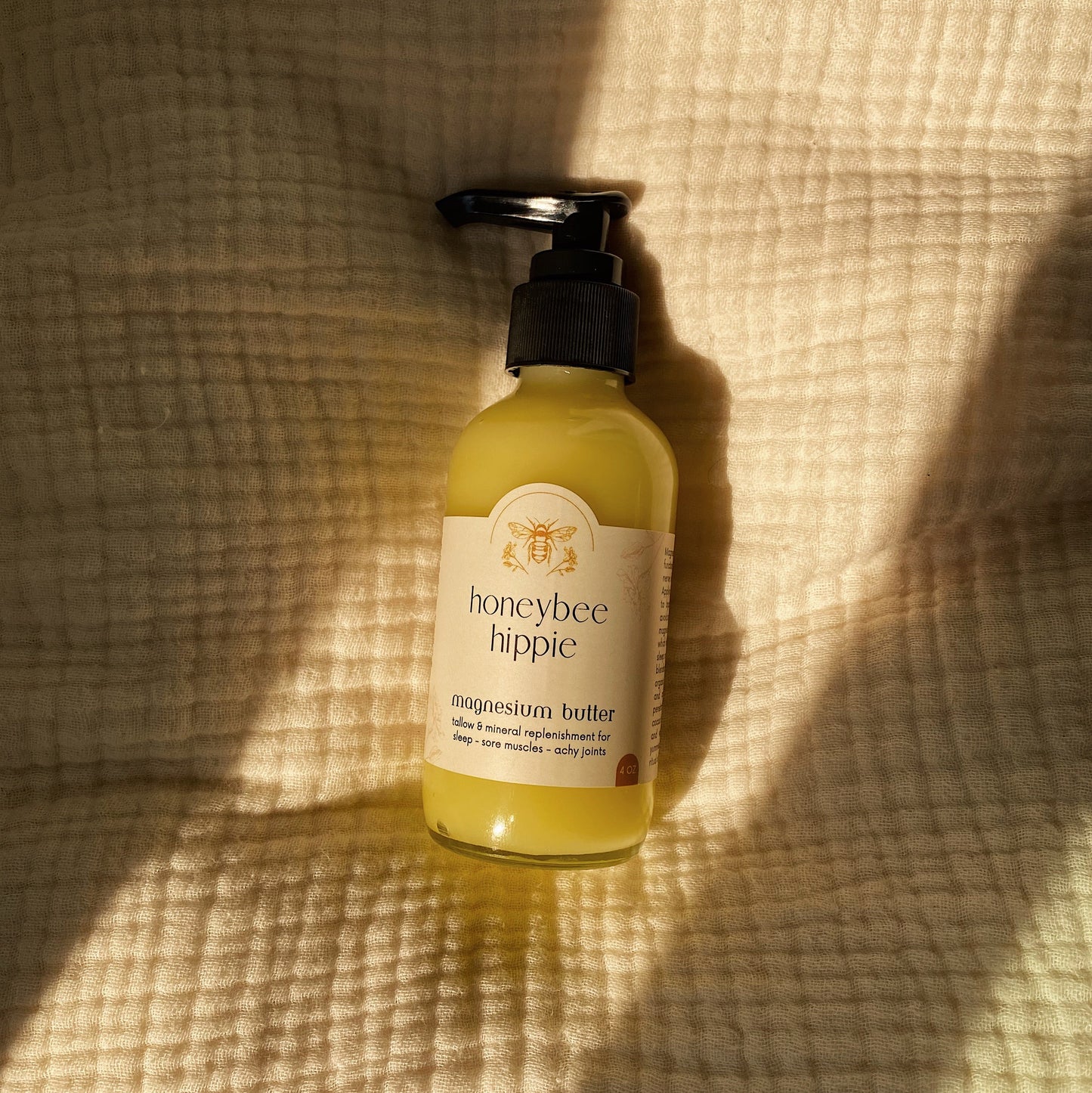 A Honeybee Hippie magnesium butter bottle with a pump rests on textured beige fabric. Sunlight creates soft shadows, inviting relaxation and promising sleep and tranquility in its gentle setting.