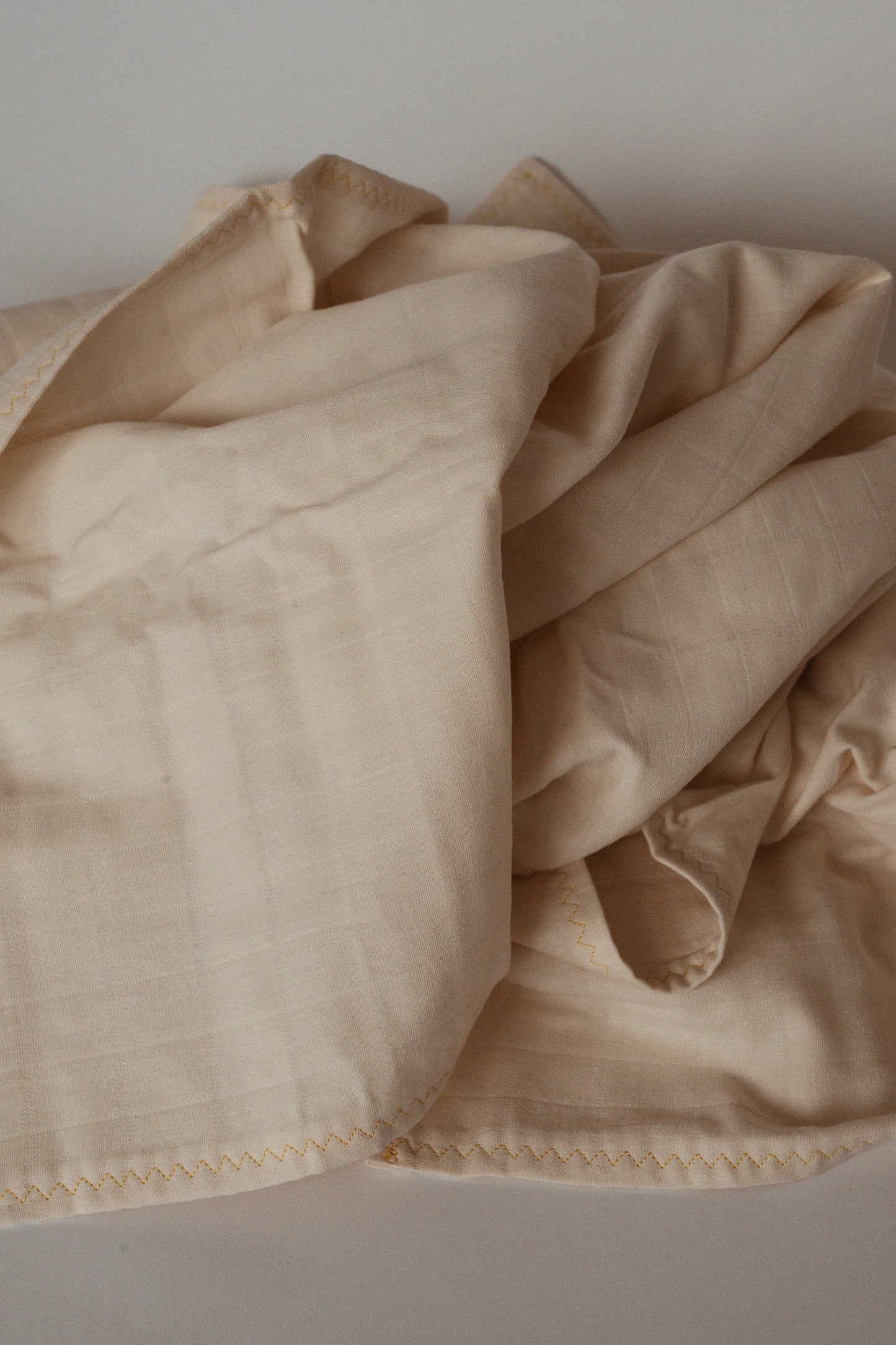 A crumpled Organic Cotton Cover by cabane childrenswear is laid on a flat surface, showcasing its visible stitching along the edges. The undyed, organic cotton provides a soft texture with small ridges that form a subtle pattern. Warm lighting accentuates the natural folds and creases of this beige cover.