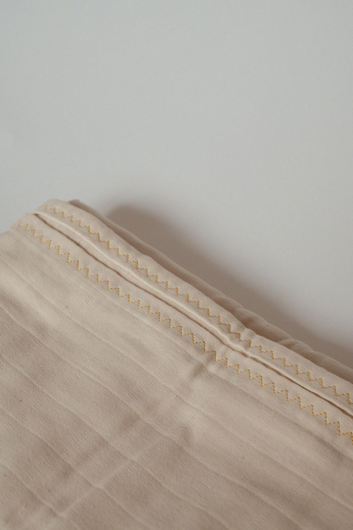 The image shows a close-up of the Organic Cotton Cover, Undyed by cabane childrenswear. This light beige, handmade piece from Vancouver features detailed stitching along its edge. The stitching pattern consists of a simple, repeating zigzag in a slightly darker shade, creating a subtle contrast against its organic background.