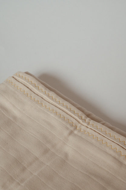 The image shows a close-up of the Organic Cotton Cover, Undyed by cabane childrenswear. This light beige, handmade piece from Vancouver features detailed stitching along its edge. The stitching pattern consists of a simple, repeating zigzag in a slightly darker shade, creating a subtle contrast against its organic background.