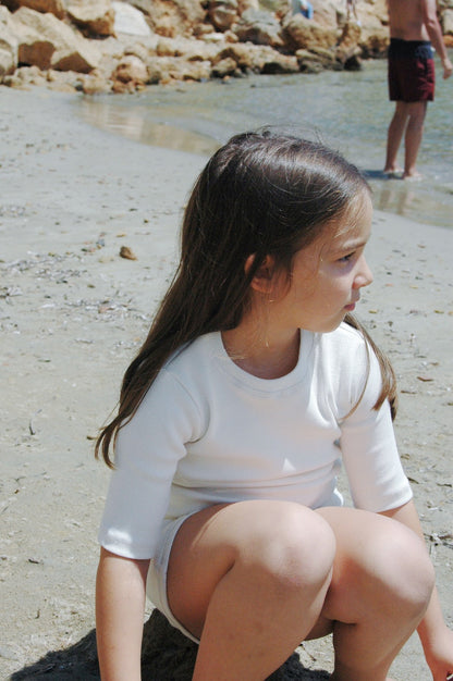 A young girl with long brown hair wearing a white outfit is crouching on the beach near the water, looking to her right. The sandy beach and rocky shoreline are visible in the background, along with another person standing in the shallow water, dressed in an ethically handmade CLASSIC SET by cabane childrenswear.