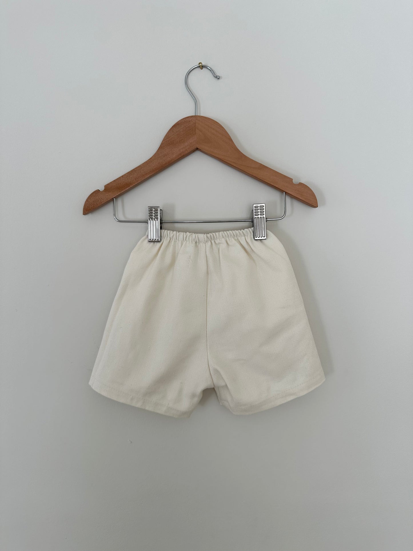 A pair of Parc Shorts in Hemp Twill from cabane childrenswear, cream-colored and made from organic cotton twill, featuring an elastic waistband, hangs on a wooden hanger against a light gray wall. The shorts showcase a simple design with no visible patterns or logos.